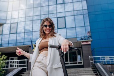 Beautiful young woman in sunglasses and white suit standing with her electric scooter near modern building and looking at camera and showing sign OK