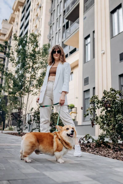 Beautiful Woman wearing white suit and sunglasses walking down the street with Welsh Corgi Pembroke dog