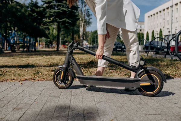 Close up of woman in white suit folding her electro scooter after ride while standing in city parkland