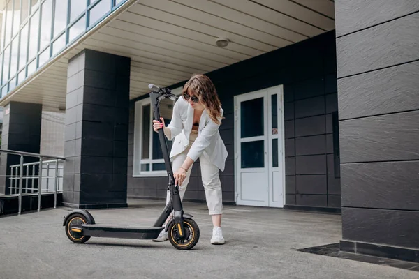 Pretty woman in white suit folding her electro scooter after ride while standing on background of modern building