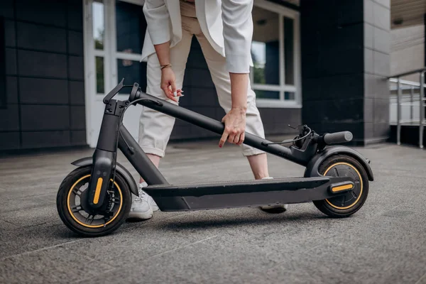 Close up of woman in white suit folding her electro scooter after ride while standing on background of modern building