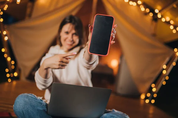 Happy Woman freelancer showing phone while working laptop on cozy glamping tent in summer night. Luxury camping tent for outdoor holiday and vacation. Lifestyle concept