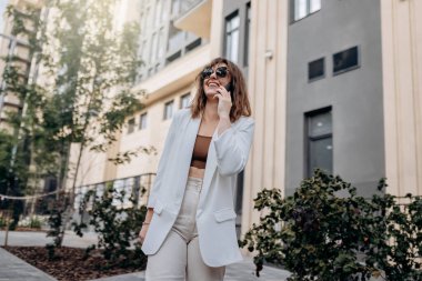 Smiling businesswoman in white suit talking phone during walking in city with modern architecture