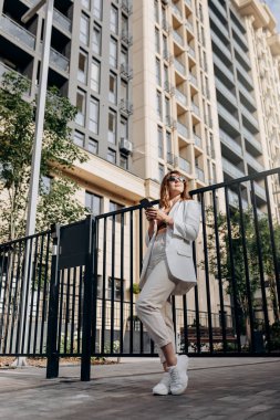Smiling businesswoman in white suit using phone during walking in city with modern architecture and looking at side