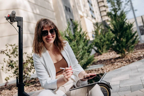 Young businesswoman in white suit sitting on electric scooter and working on digital tablet in city with modern architectury