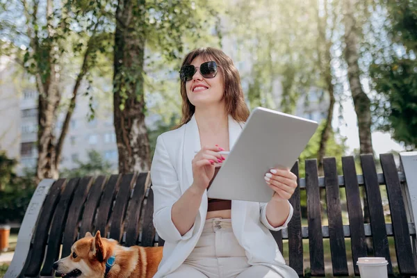 Pretty businesswoman in white suit sitting in city parkland and working on digital tablet with electric scooter on backgroun