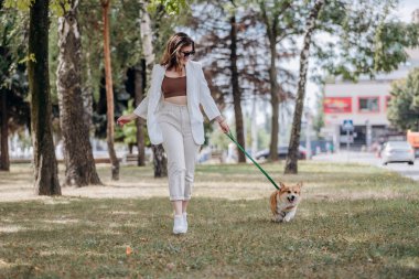 Happy Woman wearing white suit and sunglasses walking in city parkland with Welsh Corgi Pembroke dog 