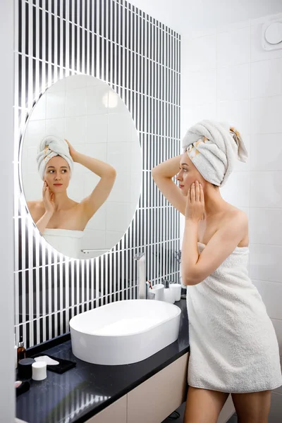 タオルの中の魅力的な若い女性がシャワーを浴びてバスルームに立ってミラーを見ています 美容とスキンケアのコンセプト — ストック写真