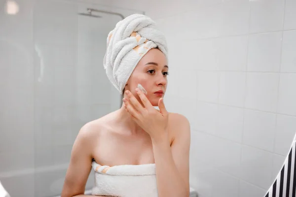 Young woman applying anti-wrinkle cream standing behind mirror in home bathroom. Cosmetology and beauty procedure. Skin care after cleansing