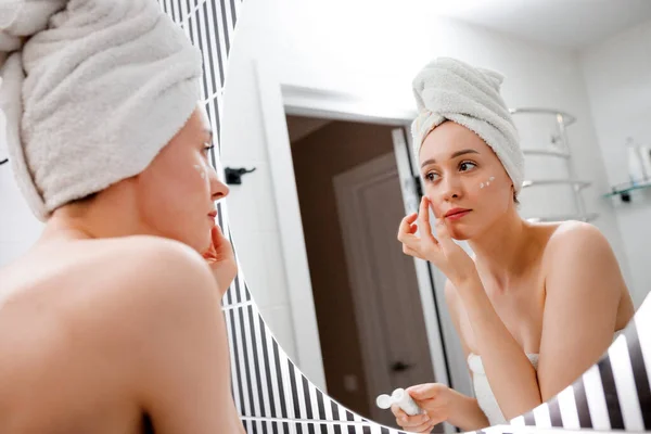 ホームバスルームで鏡の後ろに立つ抗しわクリームを適用する若い女性 美容と美容の手順 クレンジング後のスキンケア — ストック写真