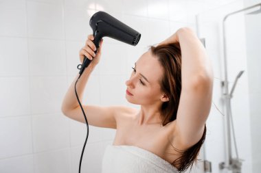Saç kurutma makinesiyle saç kurutan ve şekillendiren modern banyo içinde saç stili yapan genç bir kadın. Sabah duşundan sonra güzellik rutini