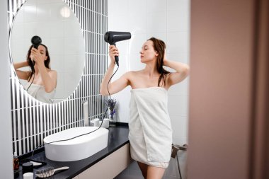 Saç kurutma makinesiyle saç kurutan ve şekillendiren modern banyo içinde saç stili yapan genç bir kadın. Sabah duşundan sonra güzellik rutini