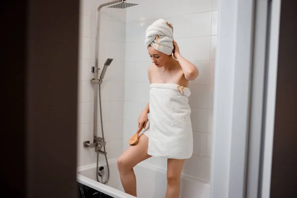 Young beautiful woman in white towel doing body lymphatic drainage massage with dry wooden brush with natural bristles in bathroom at home. Anti-cellulite exercises