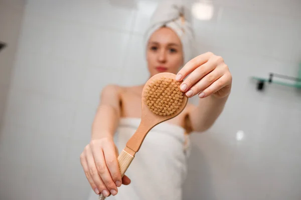 Young beautiful woman in white towel doing body lymphatic drainage massage with dry wooden brush with natural bristles in bathroom at home. Anti-cellulite exercises