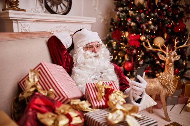 Noel Baba kanepede oturuyor ve cep telefonuyla şöminenin yanında ve Noel ağacında hediyelerle konuşuyor. Yeni yıl ve Mutlu Noeller, Mutlu Noeller konsepti