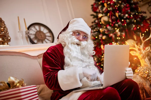 Santa Claus sitting at his home and reading email on laptop with hristmas requesting or wish list near the fireplace and tree with gifts.  New year and Merry Christmas , happy holidays concept