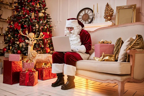 Santa Claus sitting at his home and reading email on laptop with hristmas requesting or wish list near the fireplace and tree with gifts.  New year and Merry Christmas , happy holidays concept