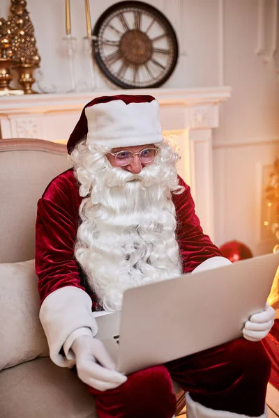 Santa Claus sitting at his home and reading email on laptop with hristmas requesting or wish list near the fireplace and tree with gifts. New year and Merry Christmas  , happy holidays concept
