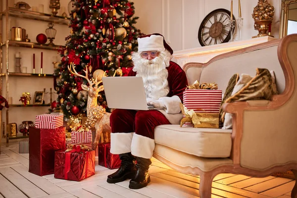 Santa Claus sitting at his home and reading email on laptop with hristmas requesting or wish list near the fireplace and tree with gifts. New year and Merry Christmas  , happy holidays concept