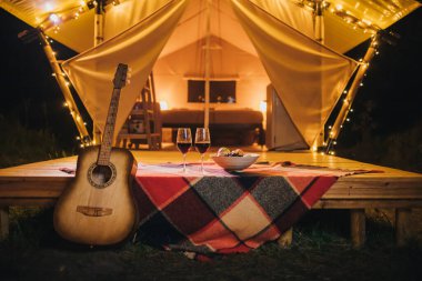 Sonbahar akşamı rahat bir çadırın arka planında duran meyveli iki kadeh kırmızı şarap ve tabak. Açık hava tatili ve tatil için lüks kamp çadırı. Yaşam tarzı kavramı