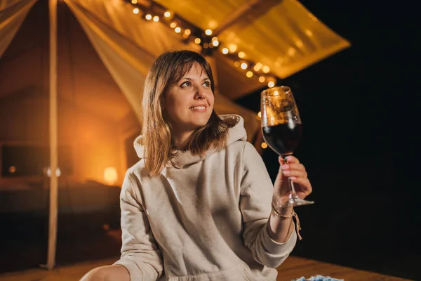 Smiling Woman freelancer drinking wine and read book sitting in cozy glamping tent in autumn evening. Luxury camping tent for outdoor holiday and vacation. Lifestyle concept