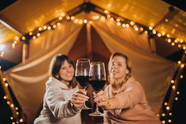 Two Smiling female friends drinking wine and eating fruits sitting in cozy glamping tent in autumn evening bonfire. Luxury camping tent for outdoor holiday and vacation. Lifestyle concept