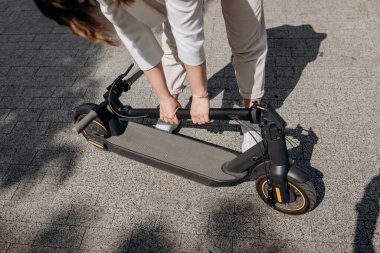 Close up of woman in white suit folding her electro scooter after ride while standing in city parkland