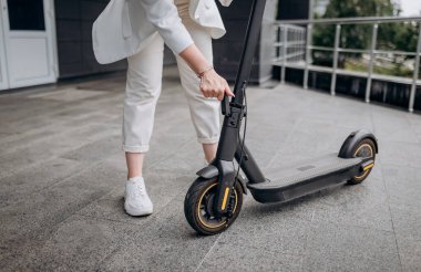 Close up of woman in white suit folding her electro scooter after ride while standing on background of modern building