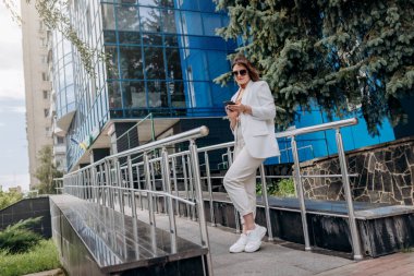 Beyaz takım elbiseli, güneş gözlüklü, gülümseyen iş kadını. Mola sırasında telefonla modern ofis binasının yanında duruyor.