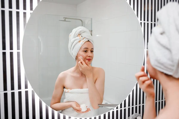 Young woman applying anti-wrinkle cream standing behind mirror in home bathroom. Cosmetology and beauty procedure. Skin care after cleansing