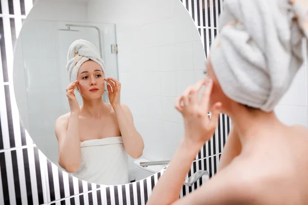 Young woman applying anti-wrinkle cream standing behind mirror in home bathroom. Cosmetology and beauty procedure. Skin care after cleansing