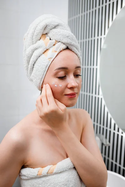 Young woman applying anti-wrinkle cream standing behind mirror in home bathroom. Cosmetology and beauty procedure. Skin care after cleansing