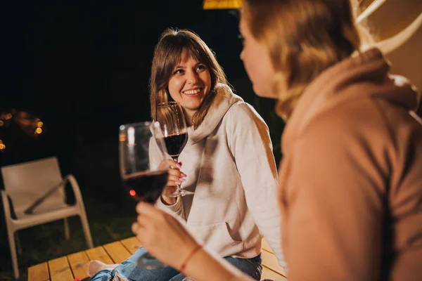 Two Smiling female friends drinking wine and eating fruits sitting in cozy glamping tent in autumn evening bonfire. Luxury camping tent for outdoor holiday and vacation. Lifestyle concept