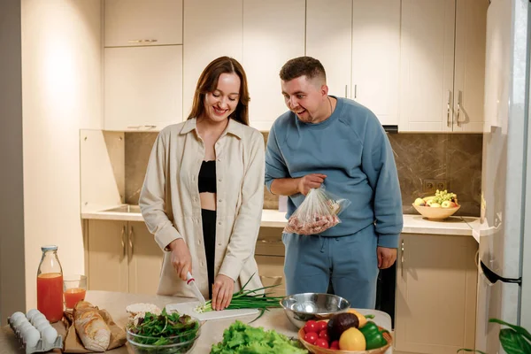 Funny man holding a bag of raw sausages and watching his wife prepare vegetable salad while standing in the kitchen at home