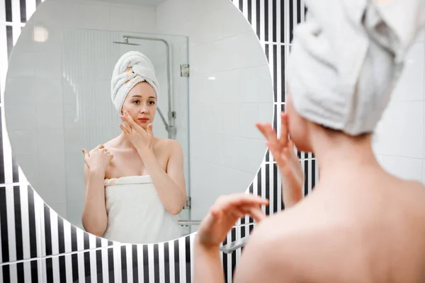 Young woman applying anti-wrinkle cream standing behind mirror in home bathroom. Cosmetology and beauty procedure. Skin care after cleansing