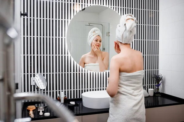 Young woman applying anti-wrinkle cream standing behind mirror in home bathroom. Cosmetology and beauty procedure. Skin care after cleansing