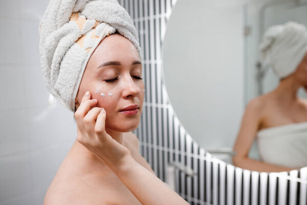 Young woman applying anti-wrinkle cream standing behind mirror in home bathroom. Cosmetology and beauty procedure. Skin care after cleansing