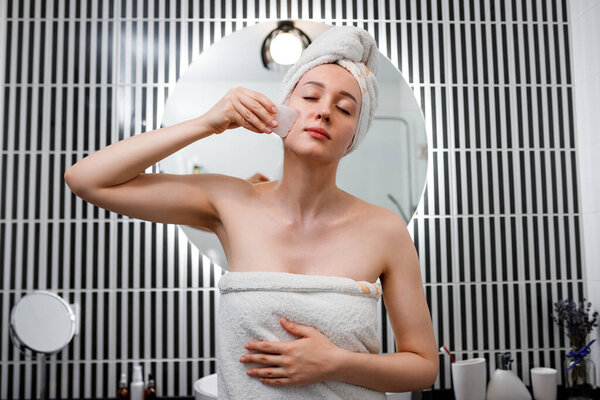 Молодая красивая женщина в белом полотенце делает массаж лица с розовым кварцевым скребком во время утренней процедуры красоты в ванной комнате. Антивозрастные процедуры и концепция skincare