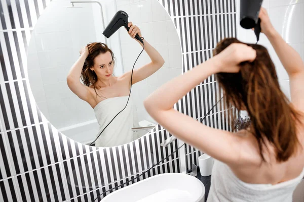 Young woman drying and styling hair with hair dryer, making hairstyle in modern bathroom interior. Beauty routine after morning shower