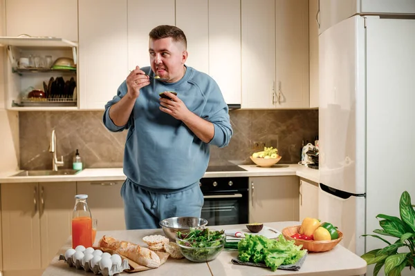 Fat and funny man eating avocado while standing in modern home kitchen. Slimming and healthy lifestyle concept