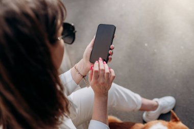 Top view of businesswoman in white suit sitting on electric scooter and using phone during walking with Welsh Corgi Pembroke dog in city