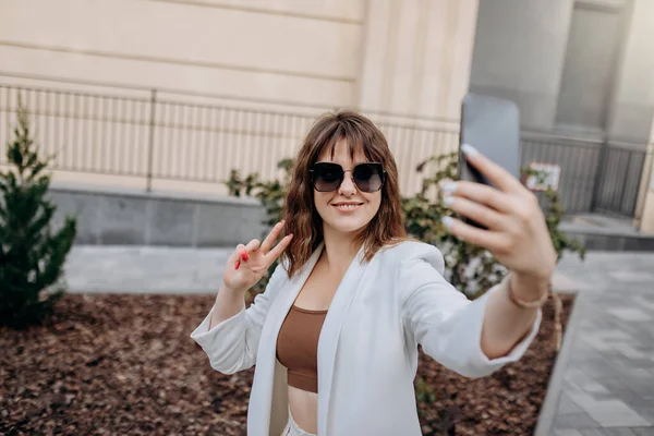 Smiling businesswoman in white suit making videocall to friends during walking in city with modern architecture