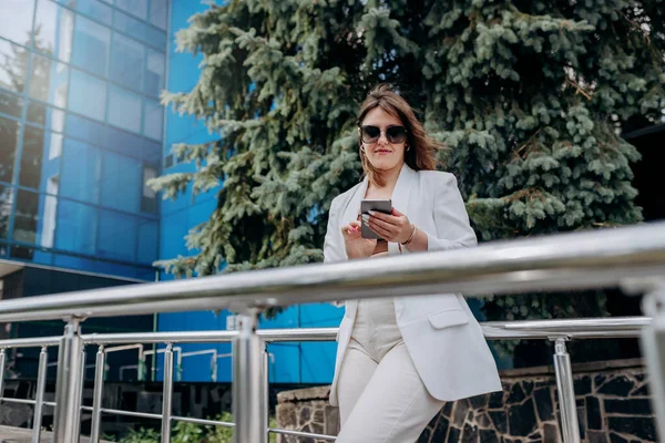 Smiling business woman in white suit and sunglasses using phone during break standing near modern office building
