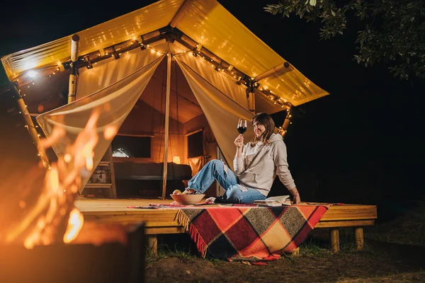 Smiling Woman freelancer drinking wine and read book sitting in cozy glamping tent in autumn evening. Luxury camping tent for outdoor holiday and vacation. Lifestyle concept