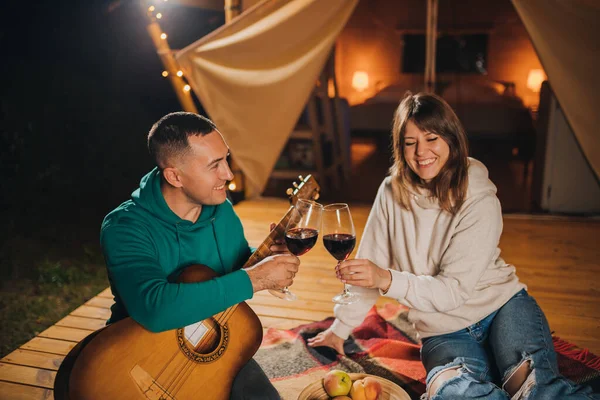 Happy couple relaxing in glamping on autumn evening, drinking wine and playing guitar near cozy bonfire. Luxury camping tent for outdoor recreation and recreation. Lifestyle concept