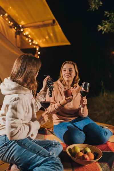 Two Smiling female friends drinking wine and eating fruits sitting in cozy glamping tent in autumn evening bonfire. Luxury camping tent for outdoor holiday and vacation. Lifestyle concept