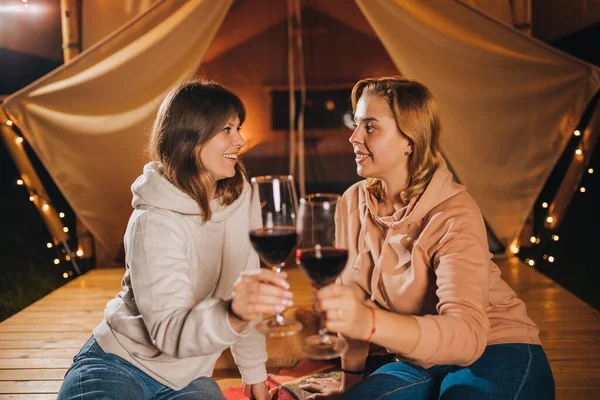 Two Smiling female friends drinking wine and eating fruits sitting in cozy glamping tent in autumn evening bonfire. Luxury camping tent for outdoor holiday and vacation. Lifestyle concept