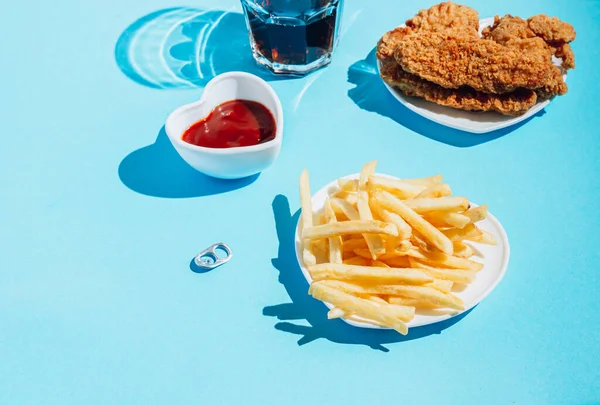 Plates in the shape of a heart with snacks on a blue background. Nuggets, wings, strips and fries with ketchup. Sunlight, harsh shadows. Selective focus with copy space