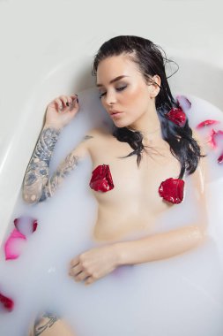 sexy brunette girl with tattoo in a bath with milk and rose petals on her nipples erotica