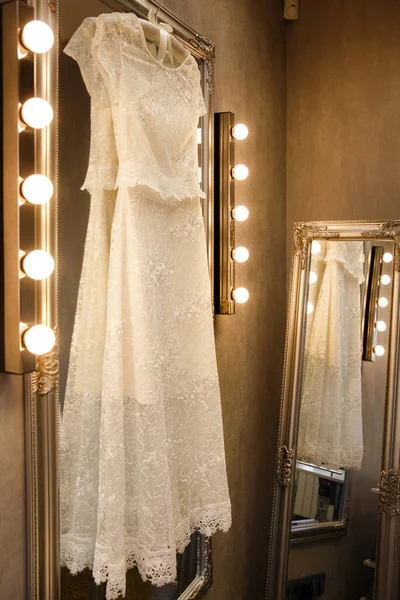 Wedding dress hanging on a mirror with lamps.